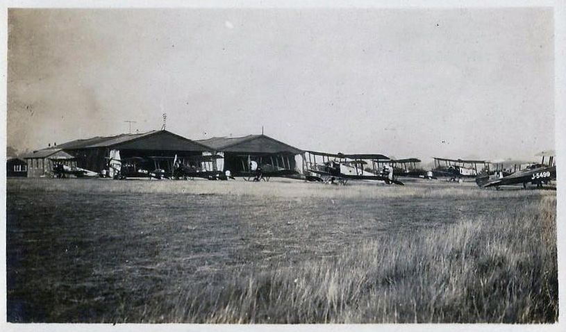 Black and whte photograph of many biplanes in front of two large hangars at an airfield.