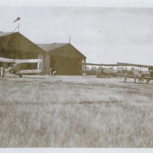Black and whte photograph of three biplanes in front of hangars at an airfield with hills in the distance.